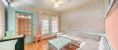 Una habitación con pared de ladrillo, ventilador de techo, ventana con cortinas, sofá, mesa y banco.
