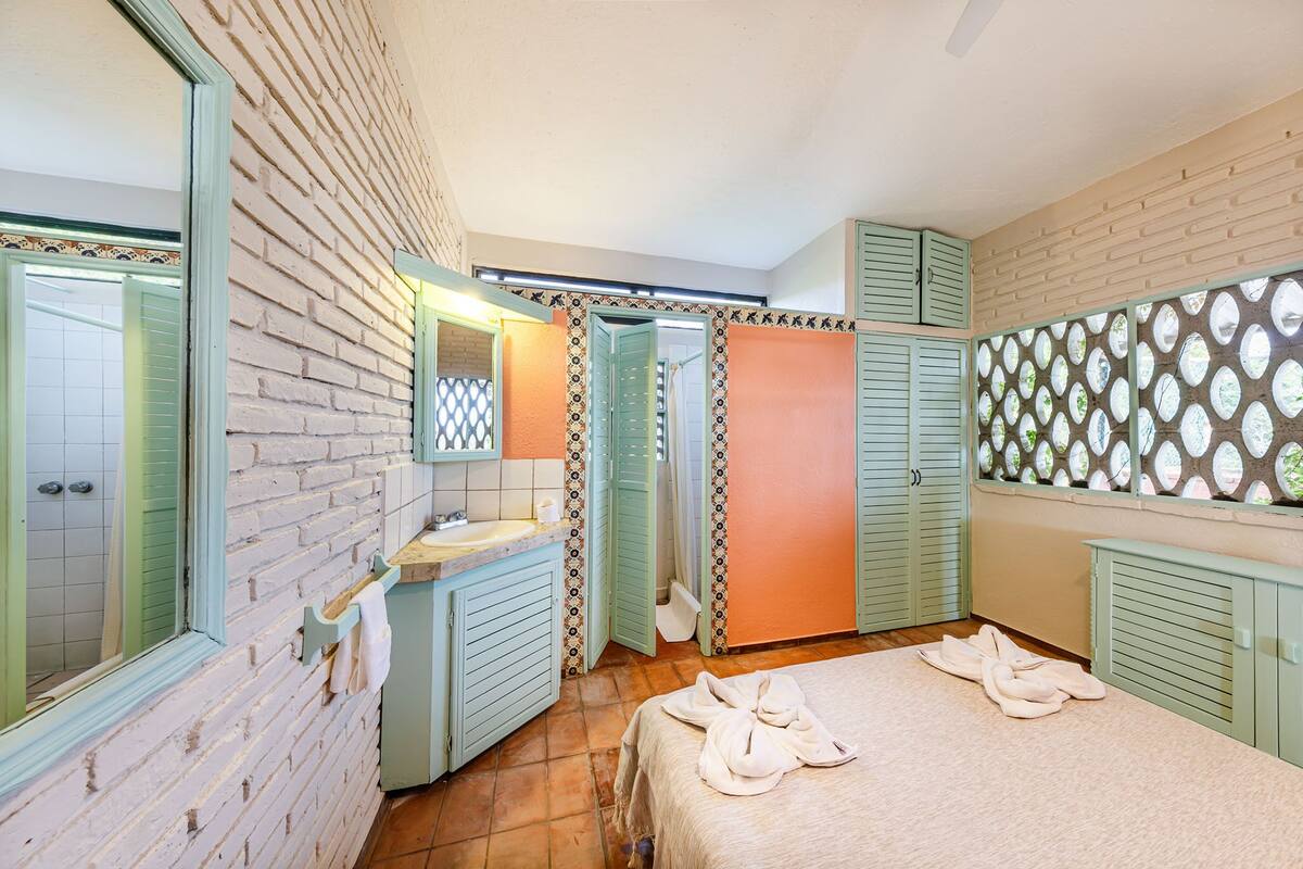 Un dormitorio con pared de ladrillo, cama con toallas, baño con lavamanos y espejo, y ventana con vista decorativa.