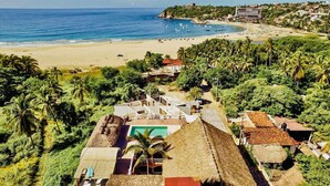 Eine Küstenlandschaft mit Strand, einem Hotel mit reetgedeckten Häusern und üppiger Vegetation.