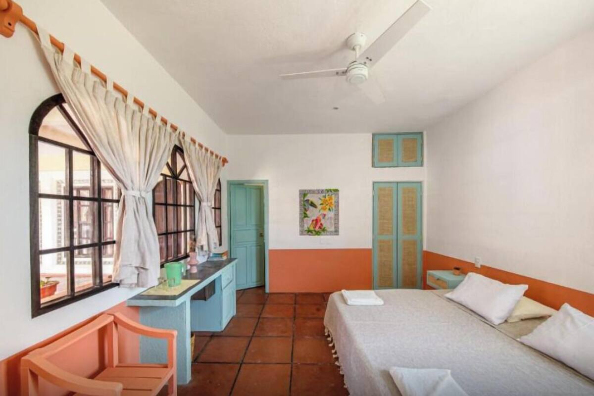 Una habitación con cama, escritorio, silla, ventana con cortinas, ventilador de techo y puerta.