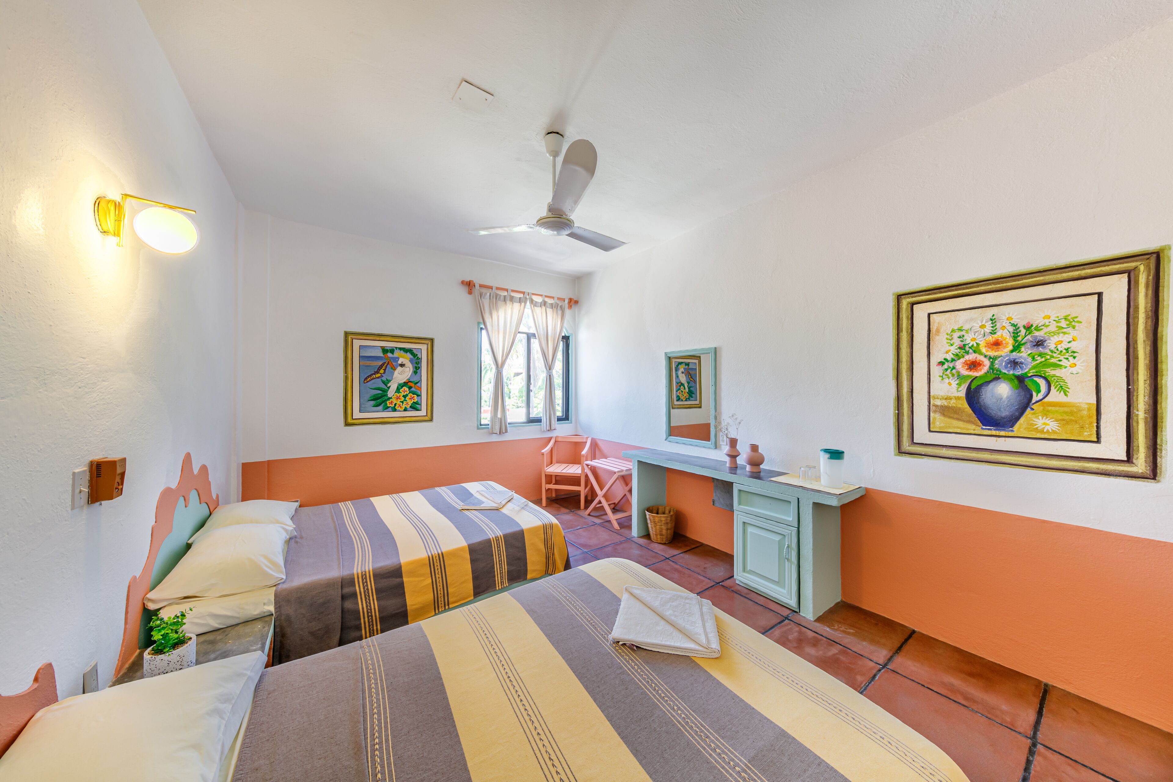 Een hotelkamer met twee bedden, een bureau, een stoel, een plafondventilator en kunstwerken aan de wanden.