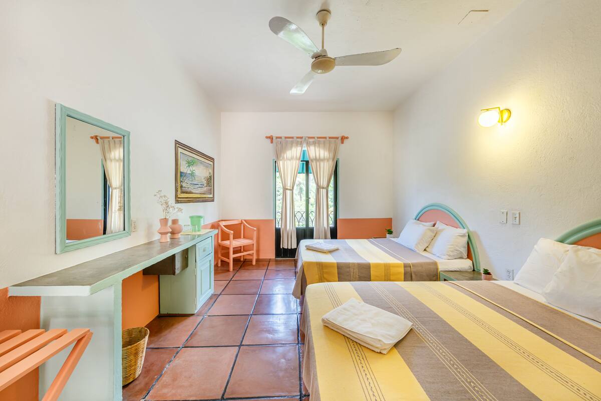 Habitación de hotel con dos camas, un escritorio, una silla, un ventilador de techo y una ventana con cortinas.