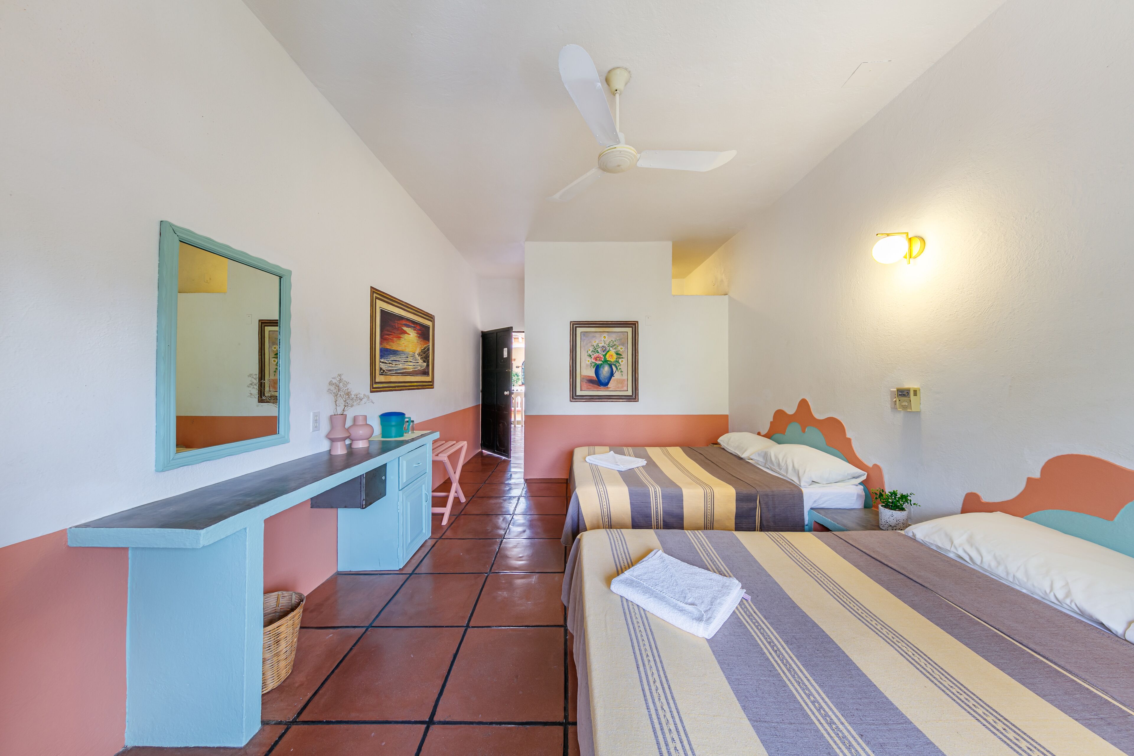 Ein Hotelzimmer mit zwei Betten, einem Schreibtisch, einem Stuhl, einem Deckenventilator und einer Wandleuchte.