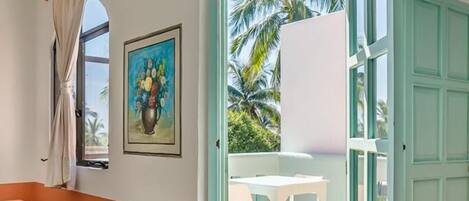 A room with a view of palm trees, a painting on the wall, and a table with chairs outside.
