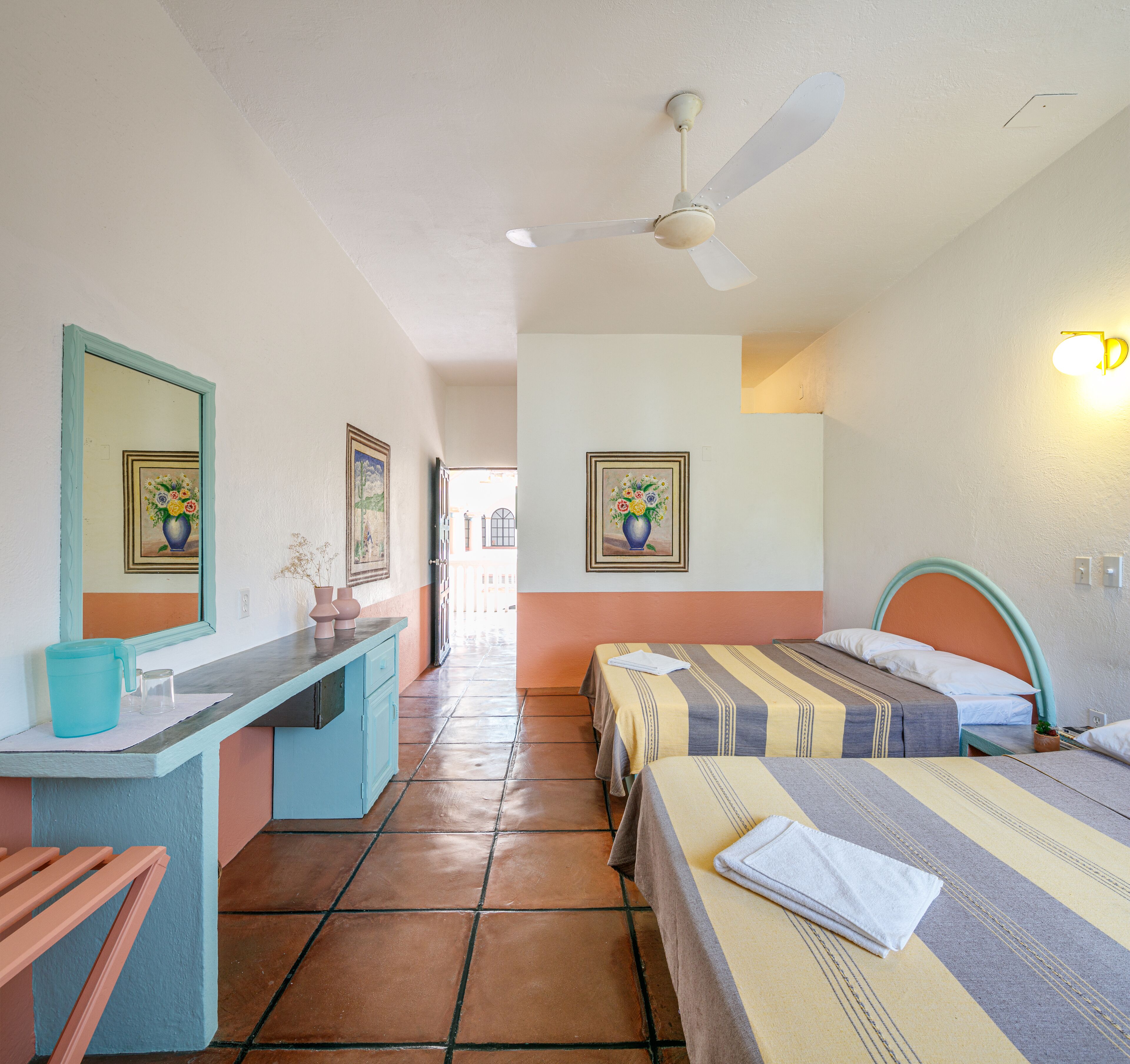 A hotel room with two beds, a desk, a ceiling fan, and artwork on the walls.
