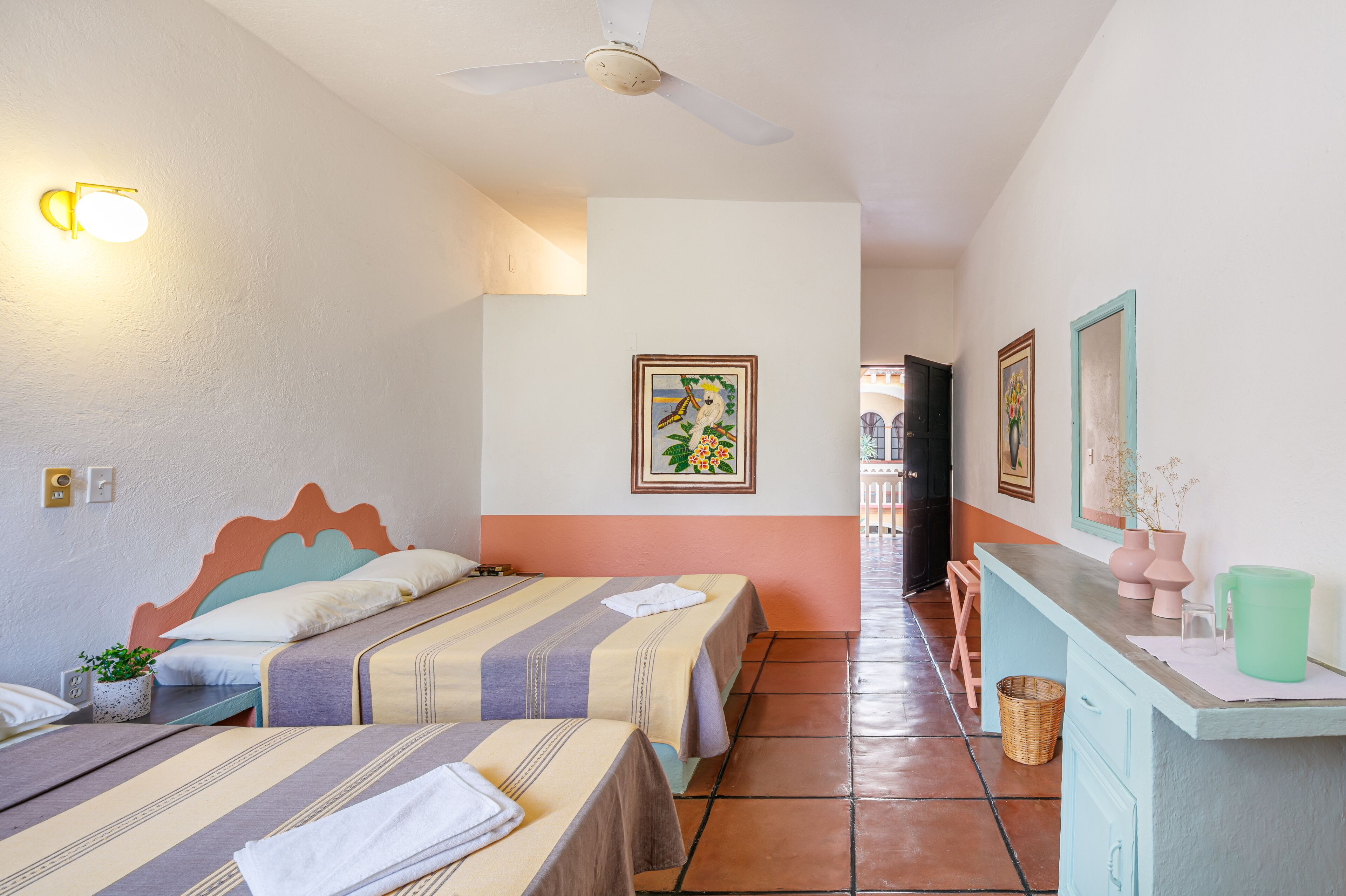 Une chambre d’hôtel avec deux lits, un ventilateur de plafond, une lampe fixée au mur, un tableau encadré, un petit bureau avec un vase et un sol carrelé.