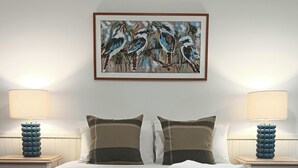 Un llit ben fet amb llençols blancs i dos coixins, flanquejat per dues taules de nit amb làmpades i gerros decoratius blaus. Sobre el llit, hi ha un quadre emmarcat amb ocells.