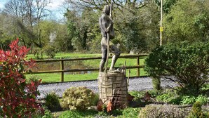 A statue of a person holding a staff, surrounded by greenery and a pond.