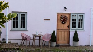 A pink building with a wooden door, two windows, a small table with chairs, and two potted plants.
