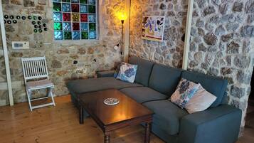 Una habitación acogedora con pared de piedra, suelo de madera, un sofá gris, una mesa de centro de madera, una silla blanca y un ventanal con vidrieras de colores.