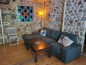 Une pièce chaleureuse avec un mur en pierre, un plancher en bois, un canapé gris, une table basse en bois, une chaise blanche et un vitrail aux couleurs vives.