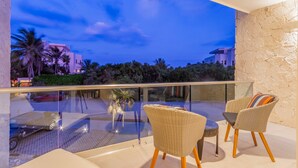 A balcony with wicker chairs and a glass table, overlooking a landscaped area with palm trees and a distant building. - Santamar by TAO (Akumal)