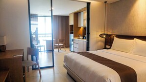 A hotel room with a bed, a desk, a chair, a kitchenette, and a view of the balcony. - Nusa Dua Suites Bali powered by Cocotel (Nusa Dua)