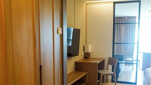 A hotel room with a wooden wardrobe, a desk with a chair, a television, and a view of the balcony. - Nusa Dua Suites Bali powered by Cocotel (Nusa Dua)