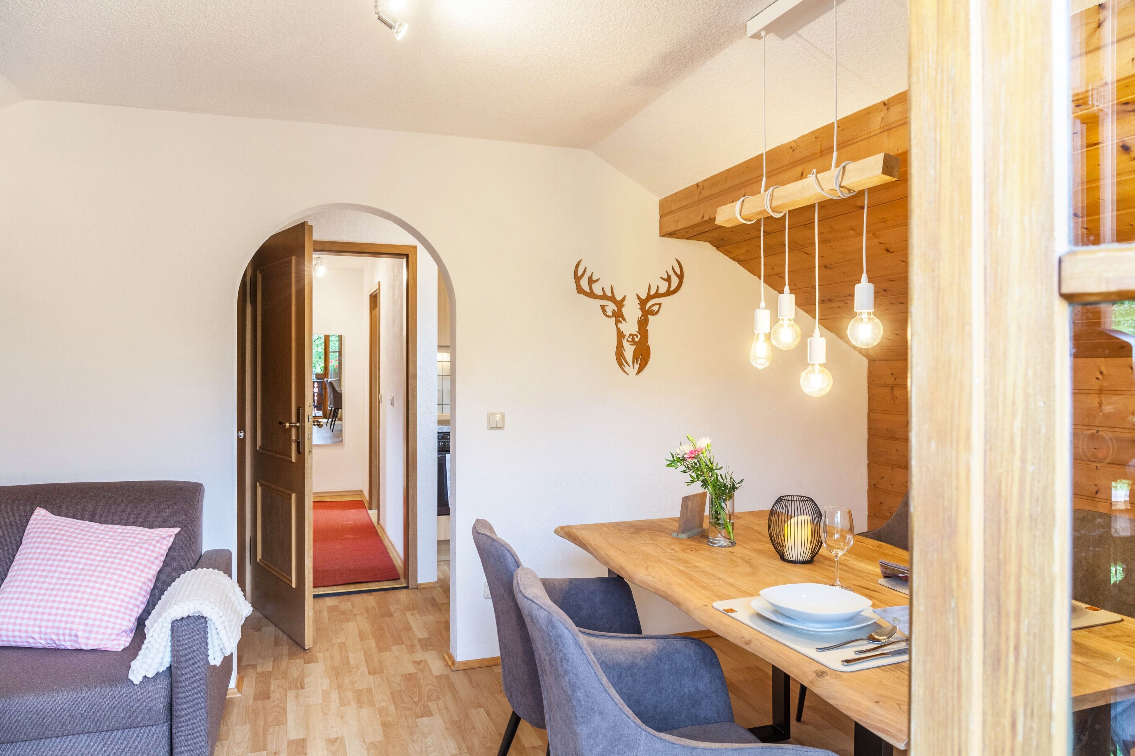 A modern living room with a sofa, dining table, and chairs. A wooden wall with a deer head decoration and hanging lights are visible.