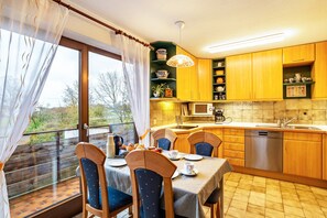 Ein Essbereich mit einem Tisch für vier Personen, Stühlen, einem Fenster mit Vorhängen sowie einer Küche mit Holzschränken und Geräten.