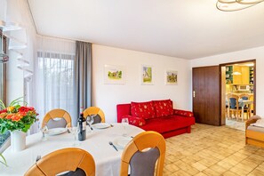 Un coin repas avec une table dressée pour deux personnes, un canapé rouge et une cuisine visible à travers une porte ouverte.