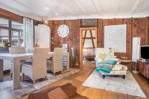 Une pièce aux murs en bois, un coin repas avec des chaises en osier, un canapé agrémenté de coussins à motifs floraux, un tapis blanc et une grande horloge au mur.