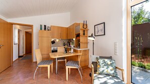 Een kleine keuken met houten kastjes, een eettafel met stoelen, een bank met een bloemenkussen, en een groot raam met uitzicht op groen.