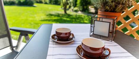 A table with two cups and saucers, a potted plant, and a view of a green lawn.