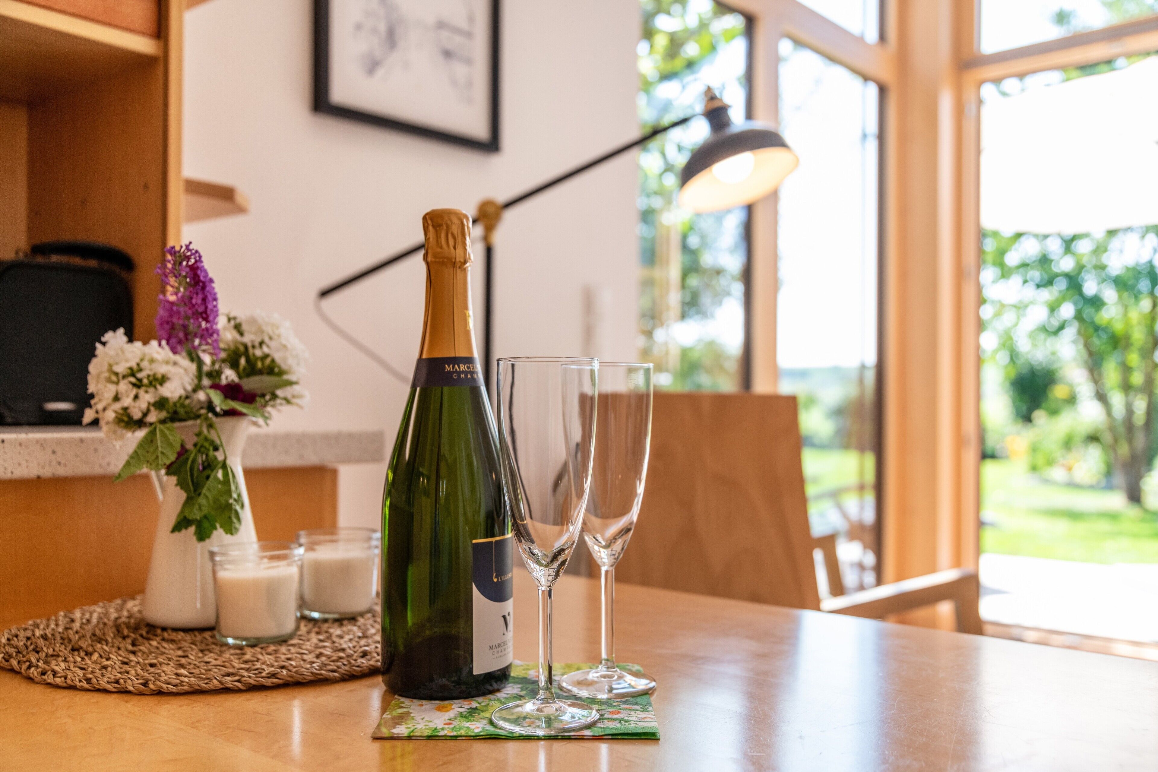 A bottle of champagne and two glasses on a table with a woven placemat, a vase with flowers, and a lamp in a room with a window and greenery outside.