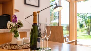 A bottle of champagne and two glasses on a table with a woven placemat, a vase with flowers, and a lamp in a room with a window and greenery outside.