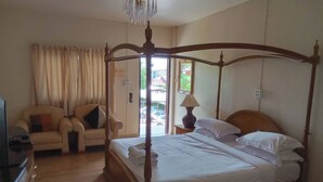 A four-poster bed with white linens, a wooden nightstand with a lamp, a sofa with cushions, and a television on a wooden cabinet. - Thai Ngam Palace Hotel (Kaeng Khlo)