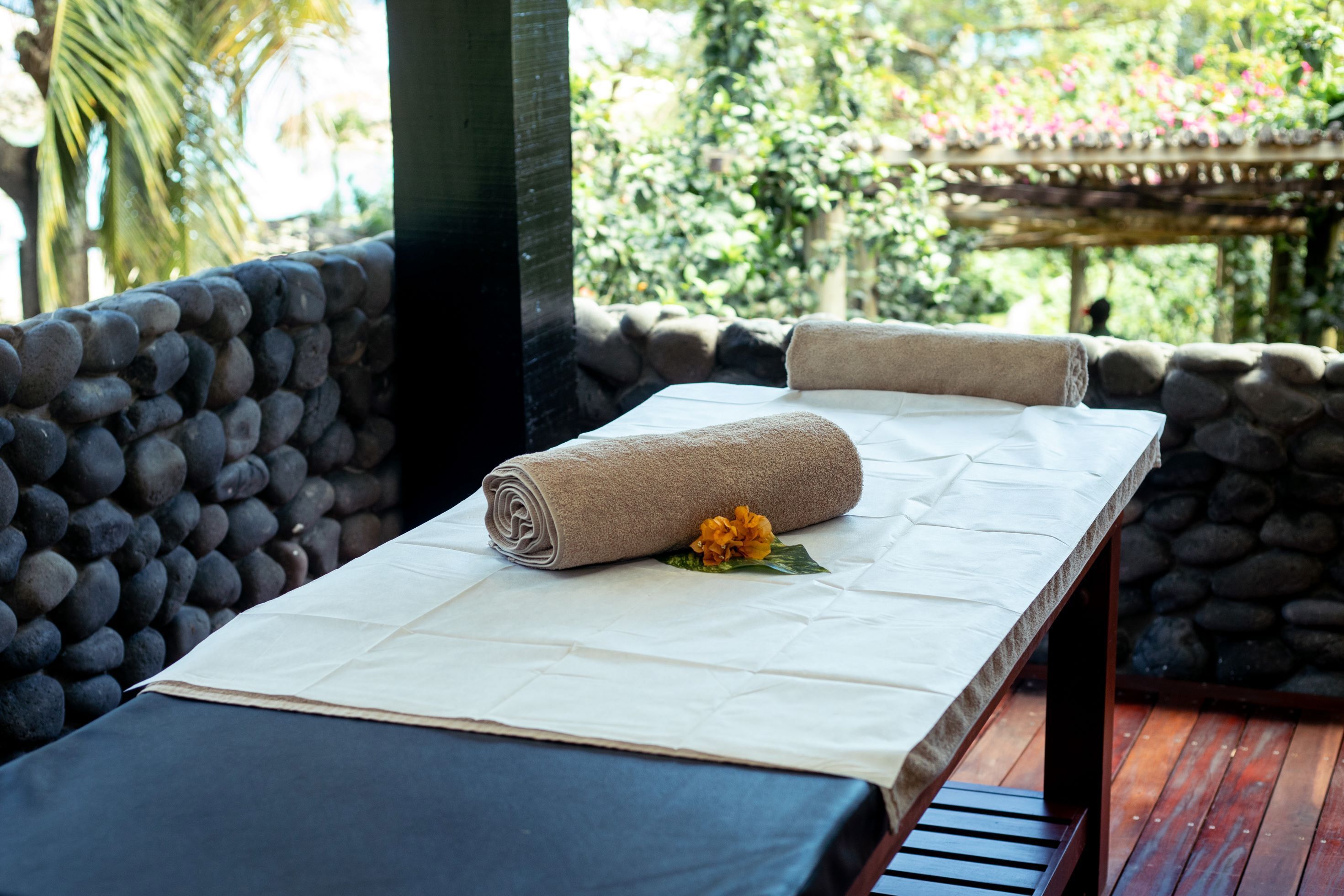A massage table with towels and a flower, set against a stone wall with a wooden deck and a garden view.