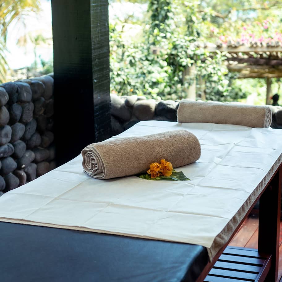 A massage table with towels and a flower, set against a stone wall with a wooden deck and a garden view.