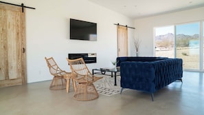 A modern living room with a large window, a blue sofa, two wicker chairs, a flat-screen TV, and a sliding door.