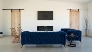 A modern living room with a large flat-screen TV mounted on the wall, two blue tufted sofas, a wicker chair, and a small round table with a vase. - New Desert Oasis | Pool, EV Charger, Spa, Short Walk to Joshua Tree Park (Joshua Tree)