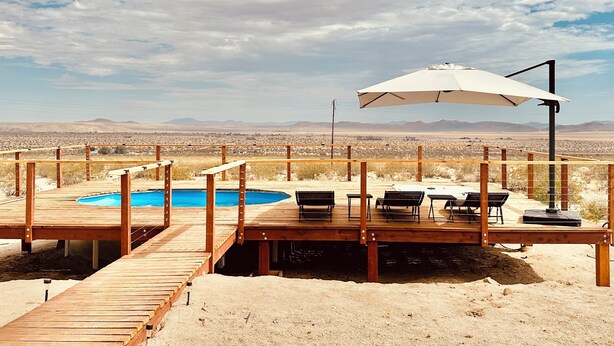 A wooden deck with a pool, outdoor seating, and a view of the desert landscape. - New Desert Oasis | Pool, EV Charger, Spa, Short Walk to Joshua Tree Park (Joshua Tree)