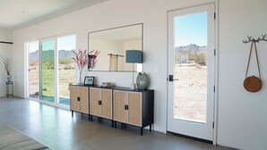 A modern interior with a large sliding glass door leading to an outdoor desert view, a black console table with decorative items, and a wall-mounted mirror. - New Desert Oasis | Pool, EV Charger, Spa, Short Walk to Joshua Tree Park (Joshua Tree)