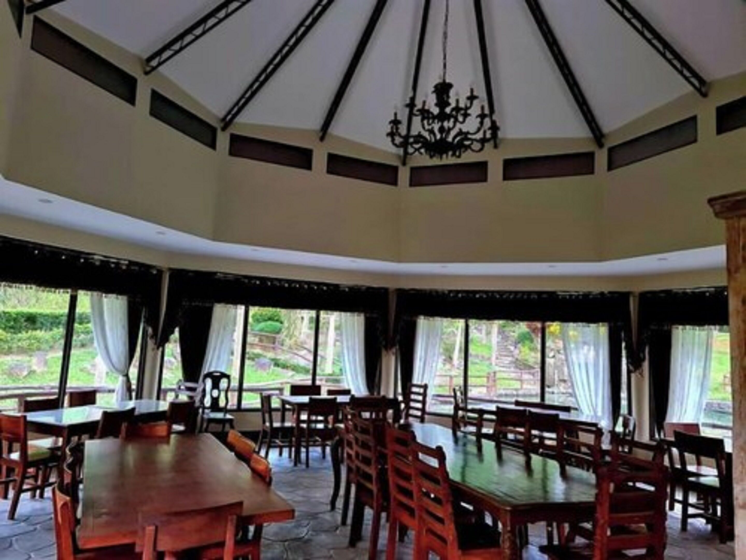 A dining area with multiple wooden tables and chairs, large windows with curtains, and a chandelier.
