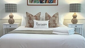 Una cama bien tendida con edredón blanco y dos almohadas decorativas, un cuadro abstracto colorido encima y dos lámparas de noche en mesitas blancas.