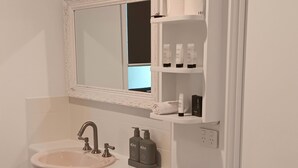 Un baño con un lavamanos blanco, dos grifos, un espejo con un marco decorativo y un inodoro. Hay artículos de aseo en los estantes y un cesto de basura.