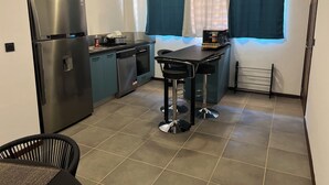 Cocina compacta con refrigerador, horno y lavamanos. Hay dos taburetes y una mesita pequeña.