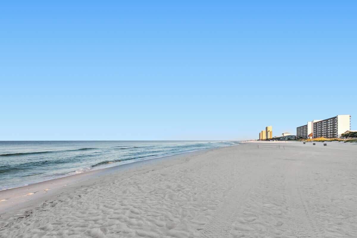 A beach with a clear sky, a sandy shore, and buildings in the background.