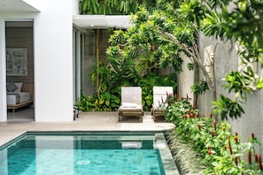 Un área de piscina con piscina azul cristalina, sillones de descanso y exuberante vegetación.