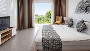Ein Hotelzimmer mit einem großen Bett, einem Fernseher auf einem Holzbalken, einem Balkon mit Vorhängen und Blick auf Grünflächen.