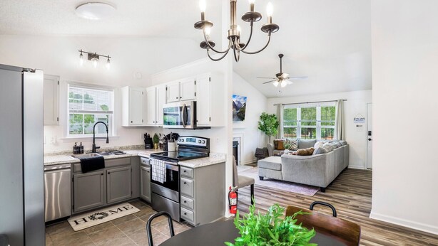 A modern kitchen with stainless steel appliances, a dining area with a wooden table, and a living room with a gray sofa and a ceiling fan. - Quaint Cottage of Holly Springs (Holly Springs)