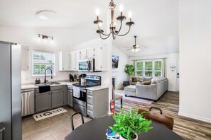A modern kitchen with stainless steel appliances, a dining area with a wooden table, and a living room with a gray sofa and a ceiling fan. - Quaint Cottage of Holly Springs (Holly Springs)