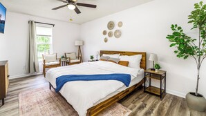 A bedroom with a wooden bed, a ceiling fan, a window with curtains, a chair, a small table with a lamp, and a potted plant. - Quaint Cottage of Holly Springs (Holly Springs)