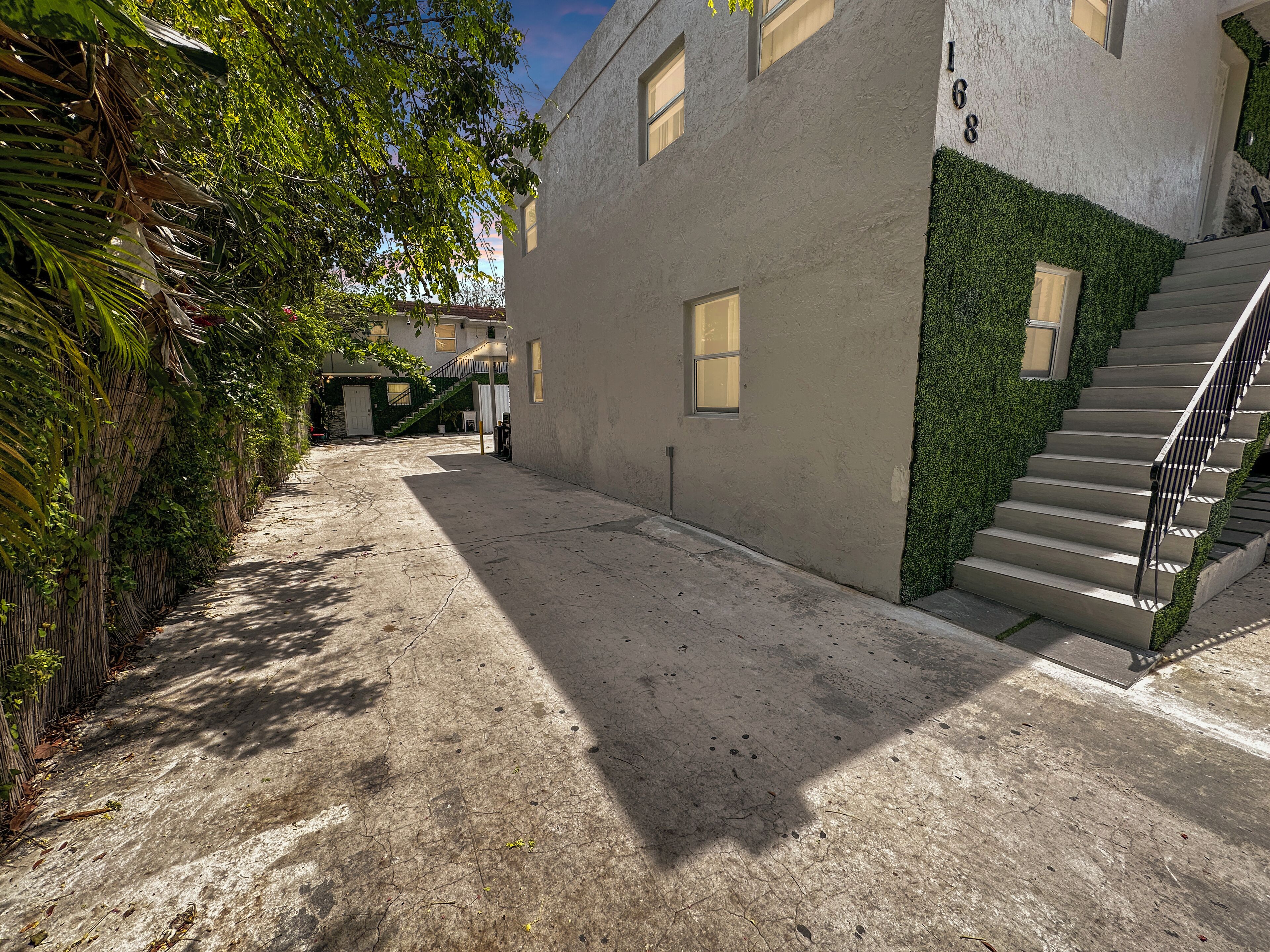 Un estrecho callejón con un sendero de hormigón, edificios adyacentes con ventanas y una escalera junto a un seto verde.
