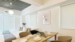 Habitación moderna y minimalista con una mesa de comedor de madera para cuatro personas, una cama con edredón gris y blanco, y una silla negra.
