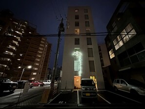 A multi-story building with illuminated signage at the entrance, surrounded by parked cars and other buildings. - New Studio Apt in the City Center for 6Ppl (Naka Ward, Hiroshima)