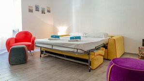 Una habitación de hotel pequeña con una cama, una silla roja, una mesita y un reposapiés morado.
