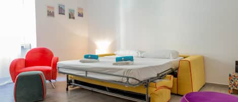 Una habitación de hotel pequeña con una cama, una silla roja, una mesita y un reposapiés morado.