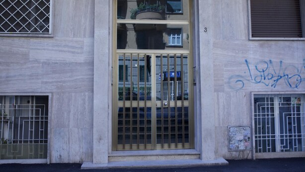 Entrada de un edificio con una puerta protegida por barrotes, un pequeño balcón y un muro con grafitis.