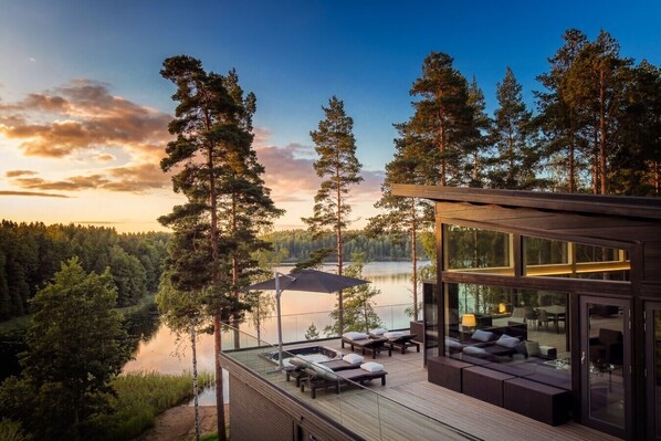 A modern cabin with a deck overlooking a lake, surrounded by tall trees.